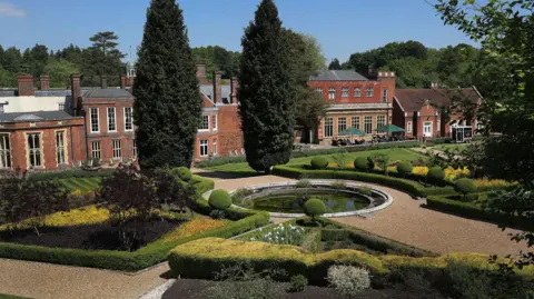 Wotton House Hotel A gravel path leads to and around a water feature in a manicured garden, with carefully pruned hedges and trees. The path leads to a large brick manor house.