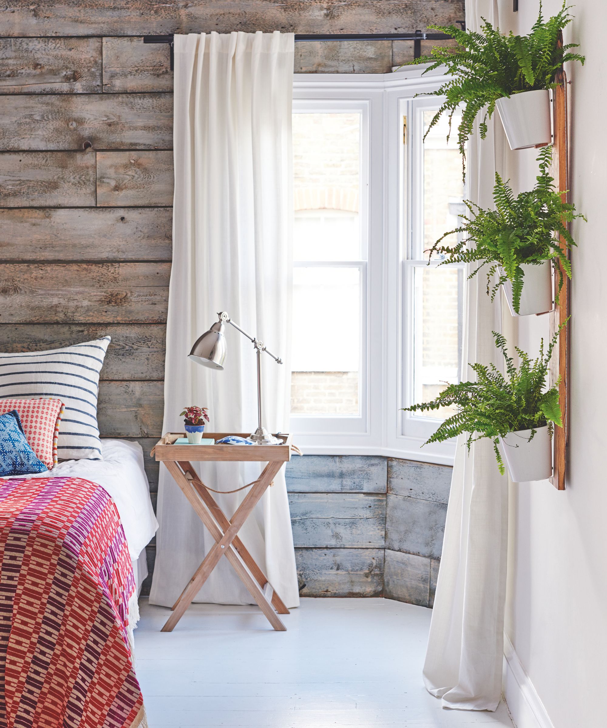 An airy wood panelled bedroom with white curtains, houseplants, and colorful patterned bedding.