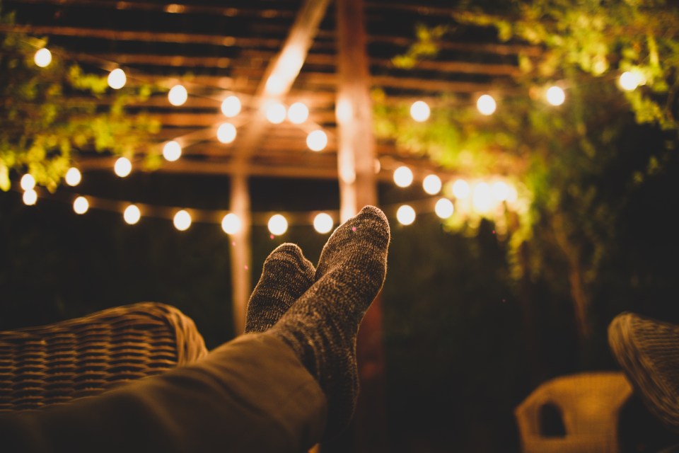 Person in socks relaxing on a patio at dusk, with string lights overhead.