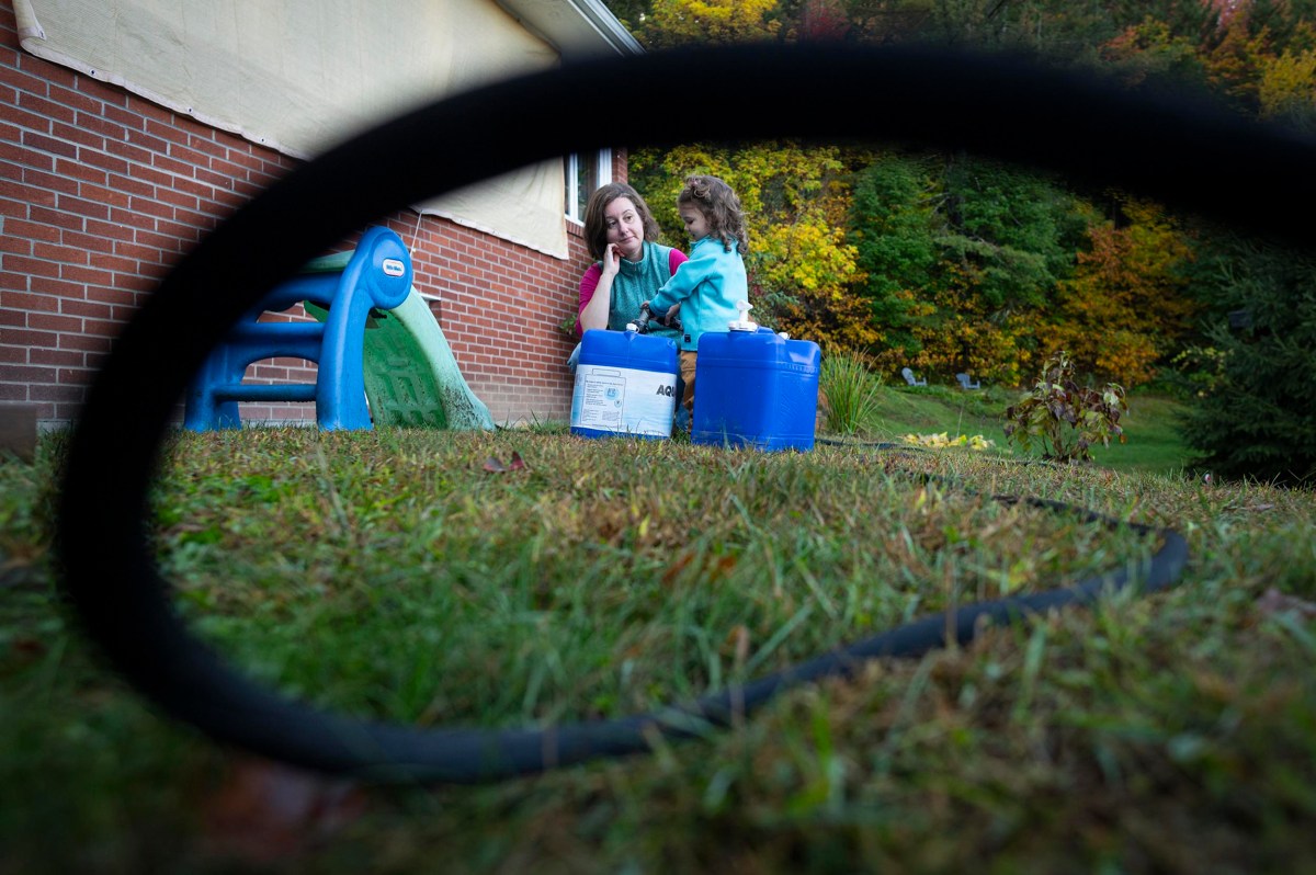 A woman and a child sit outside near a brick house, with blue containers beside them; a black hose curves in the foreground, and trees with autumn foliage are in the background.