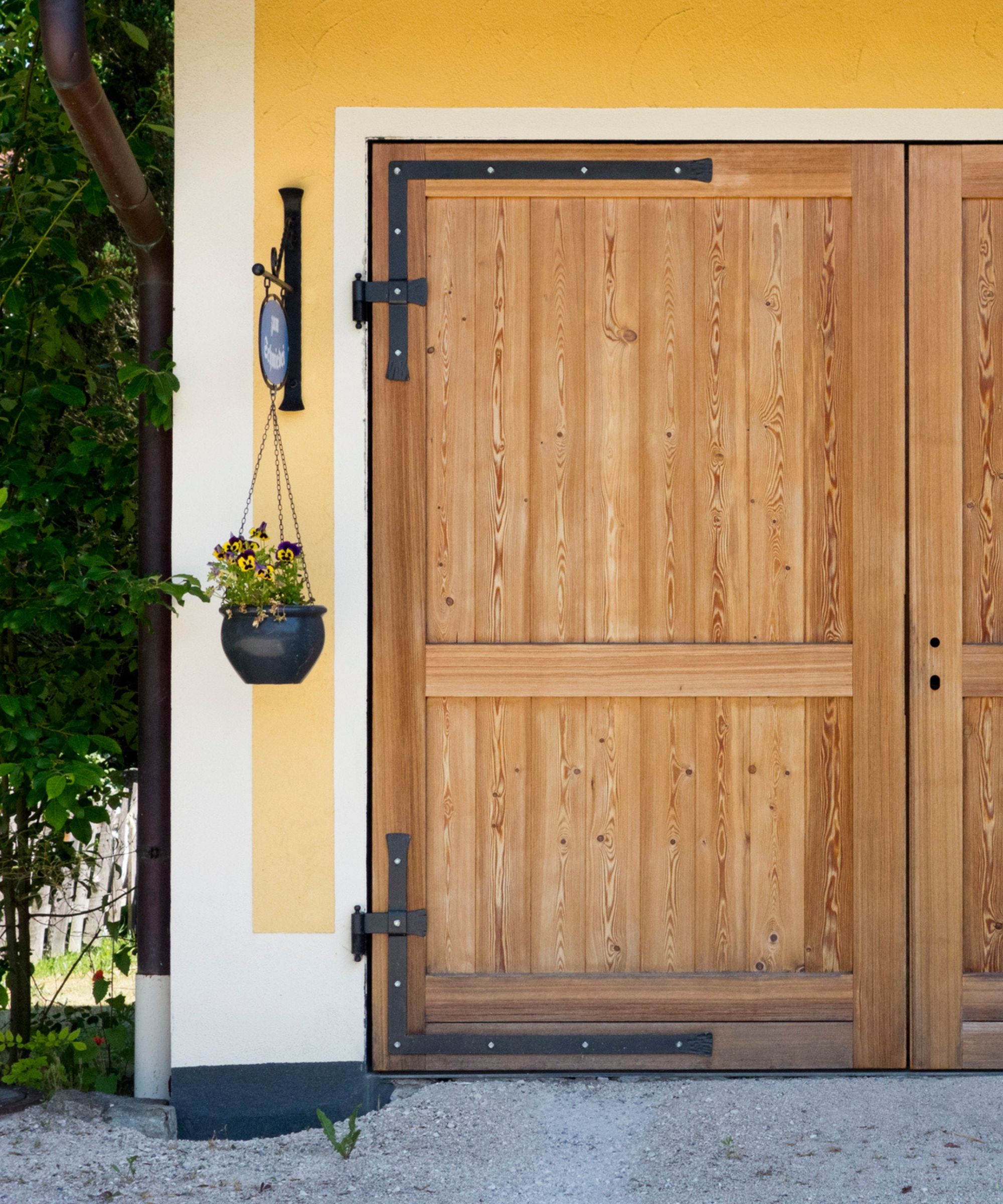 A wooden garage door with a yellow border around it, a hanging basket to the left, and gray gravel in front of it