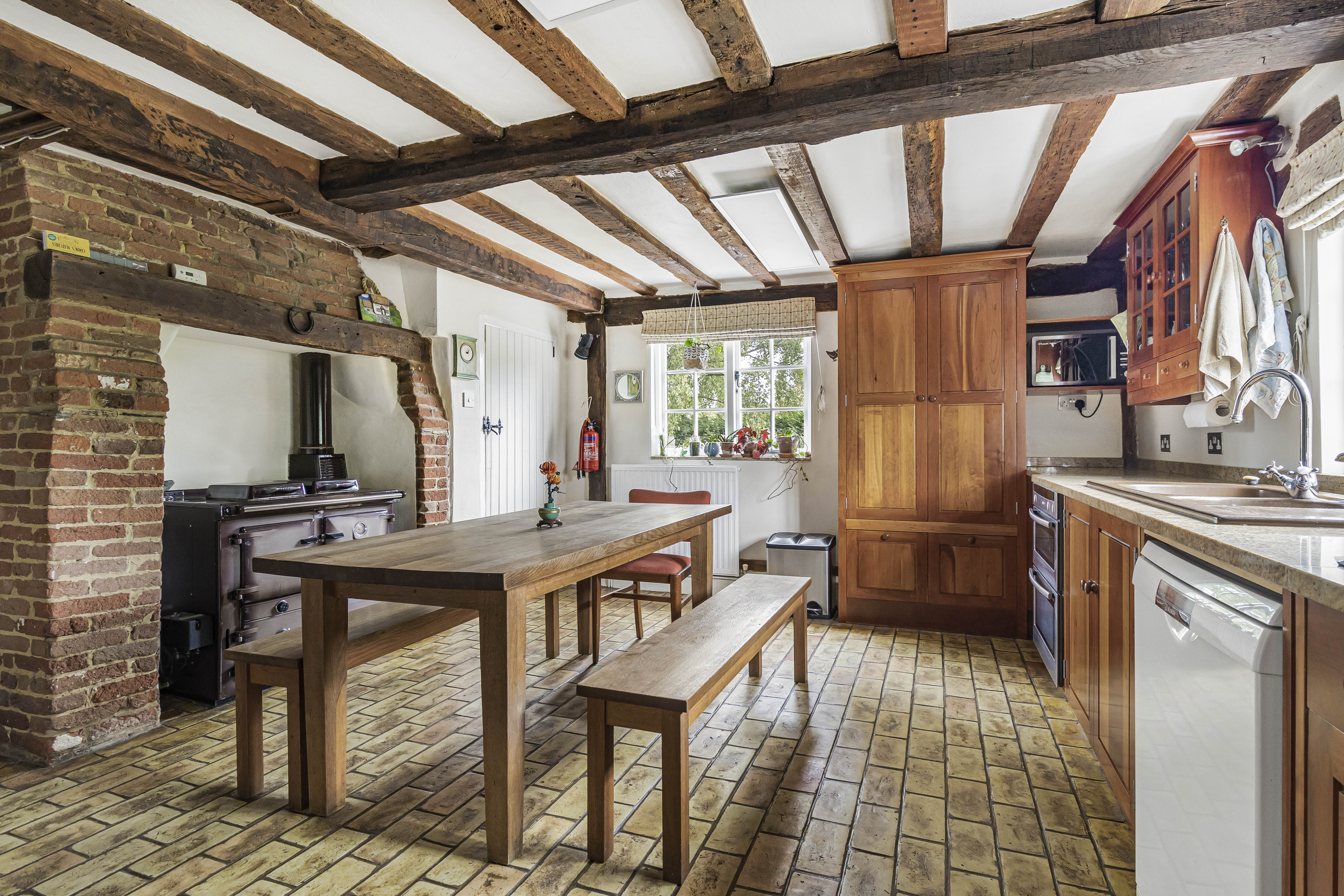A rustic kitchen with a long wooden table, benches, and a chair on terracotta tile flooring.