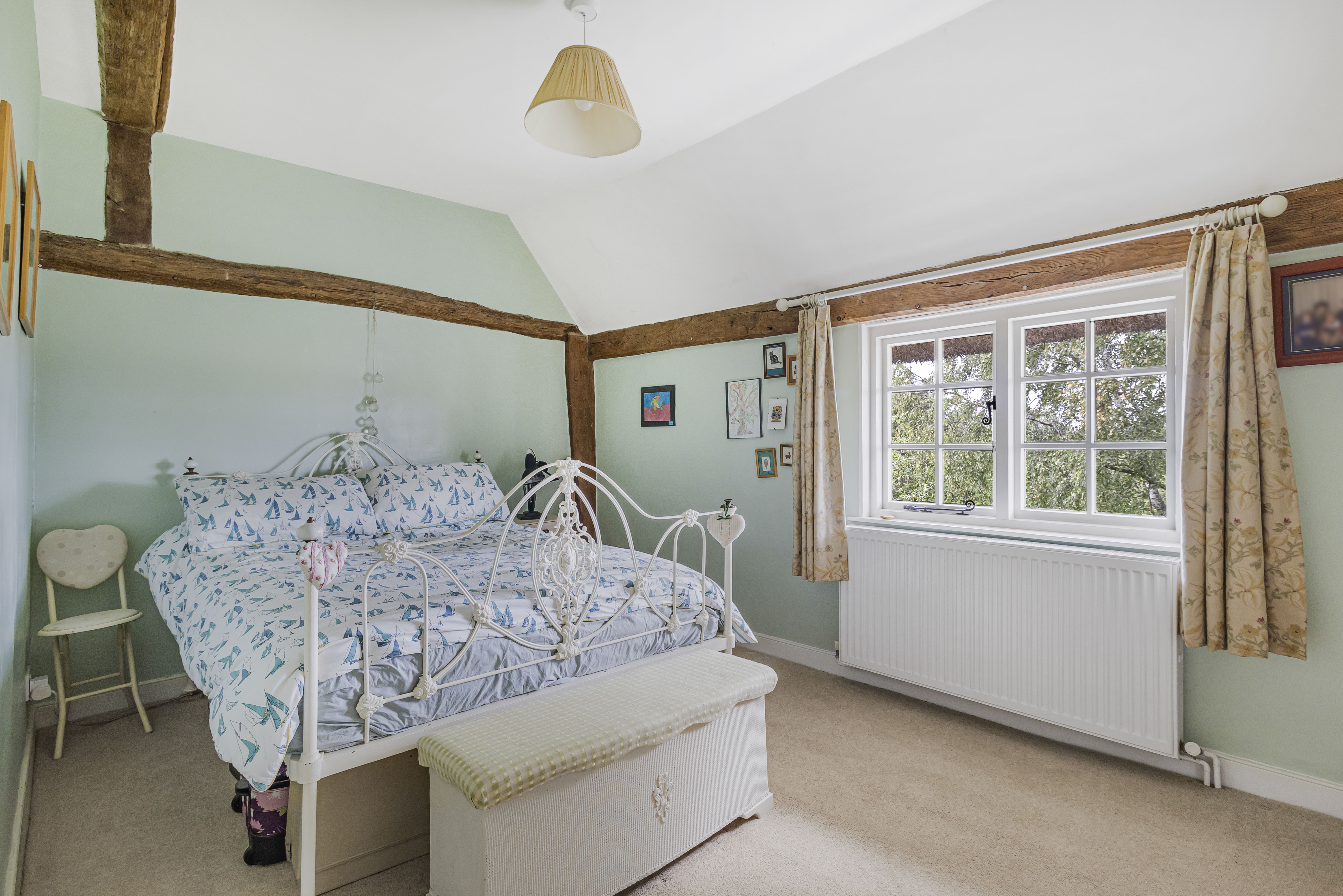 Bedroom with a white iron bed frame, a white woven chest at the foot of the bed, and pale green walls with exposed wooden beams.
