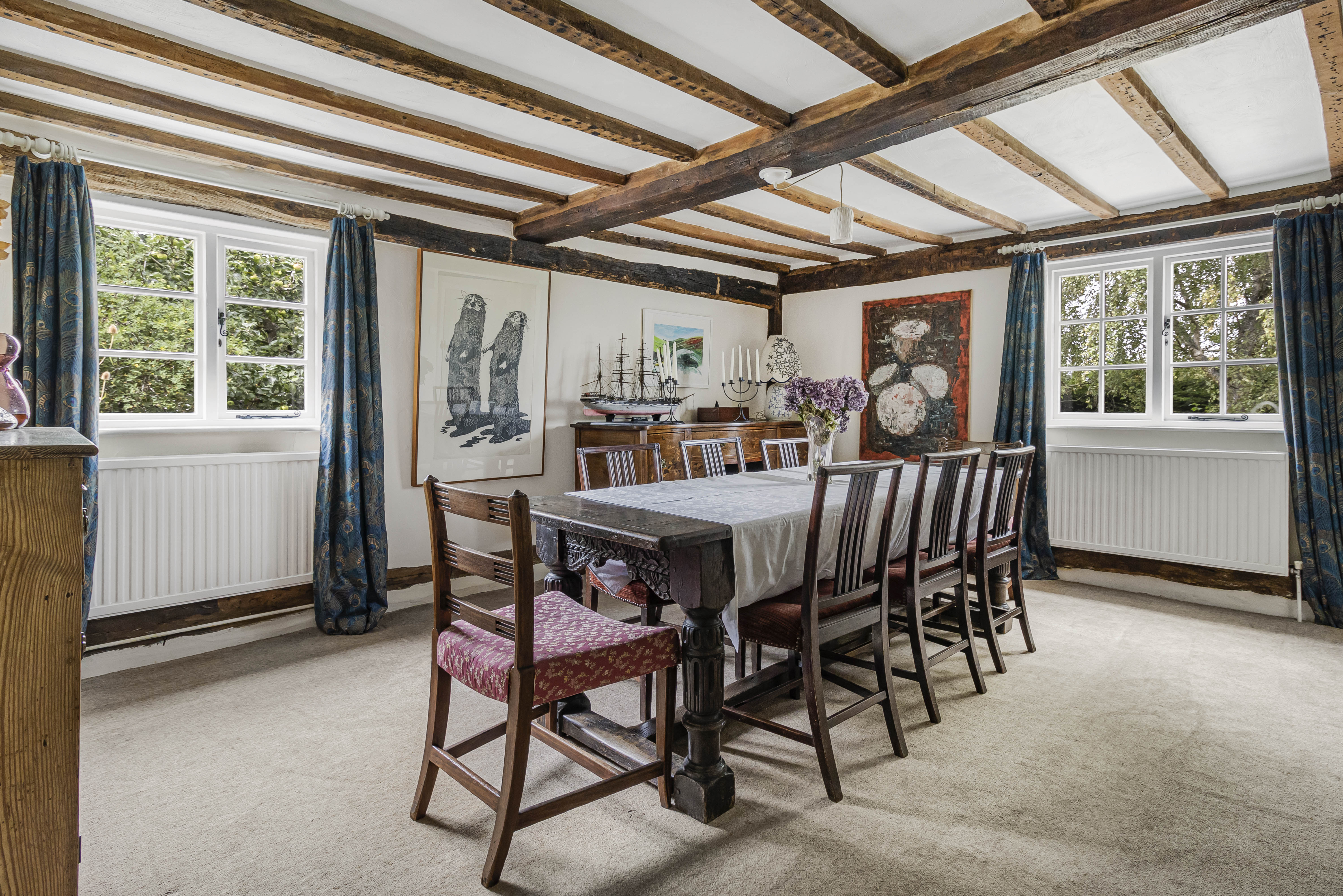 A dining room with dark wooden furniture, exposed ceiling beams, and blue patterned curtains.