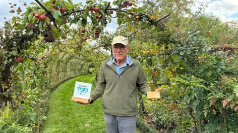 Wendy Hurrell/BBC Steve Mills holding his two trophies, standing underneath his fruit tree arched tunnel. Apples hang down above his head.