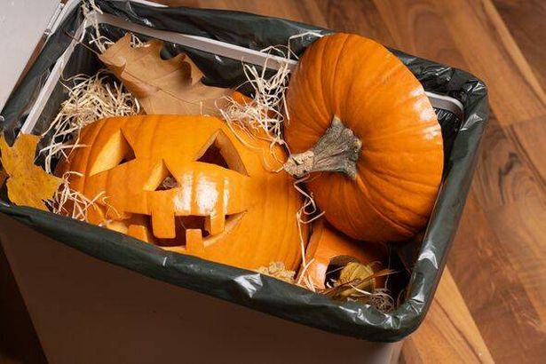 Orange pumpkins, one carved with a spooky face, are placed in a trash bin surrounded by autumn leaves and straw, symbolizing Halloween festivities and