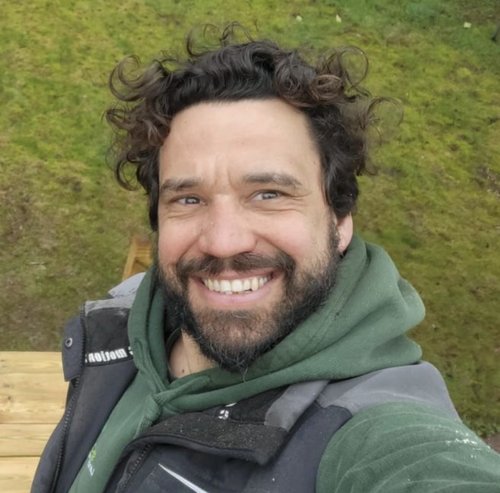 A man with curly hair and a beard smiling at the camera, wearing a green hoodie and a dark jacket, with green grass in the background.