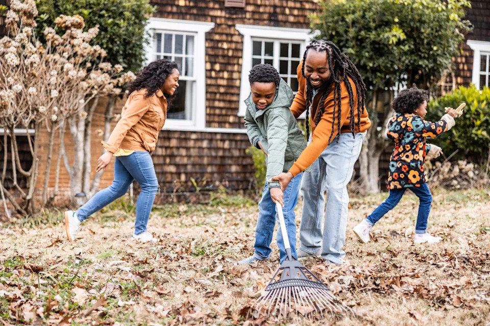 Father teaching son to rake leaves while family is playing in suburban yard