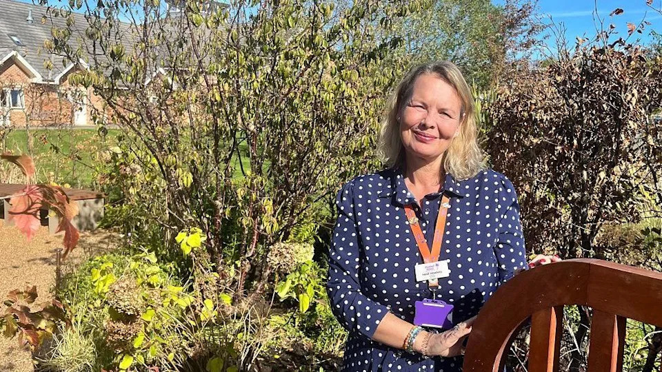 A woman with blonde hair stands in a garden next to a brown fence. She is wearing blue and white spotty dress, and has a lanyard round her neck. 