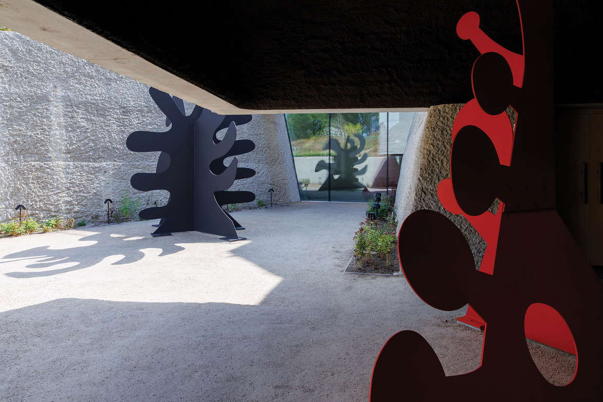 Outdoor art installation with red and black abstract sculptures casting shadows on a sunlit path between textured walls.