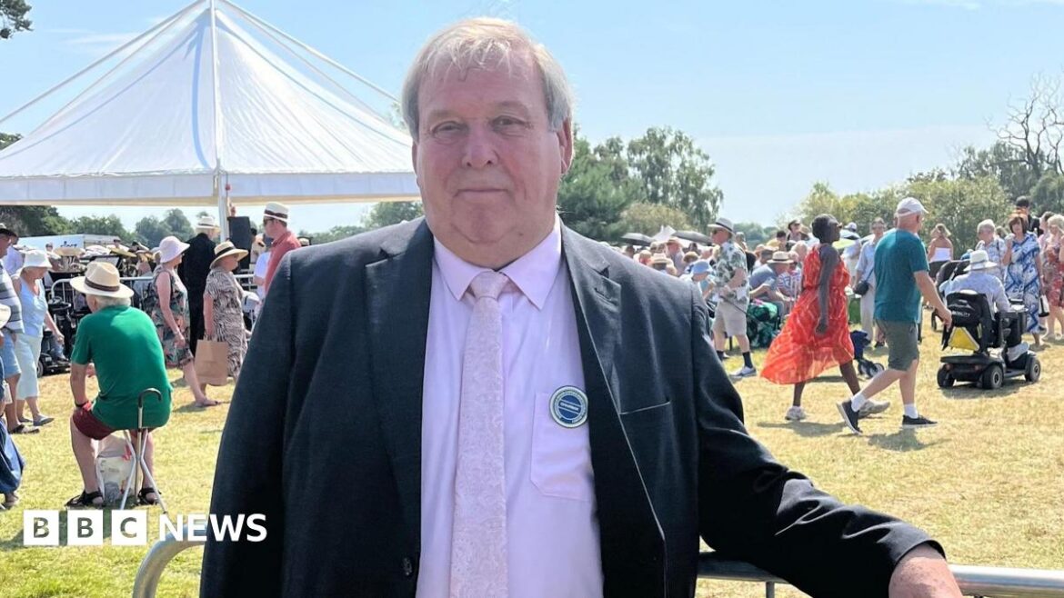 Ticket prices nearly double for RHS Sandringham Flower Show Graham Browne looks directly at the camera as he is photographed outside with crowds of people behind him. He is wearing a dark suit with a pink shirt and pink tie. There is a open marquee behind him.