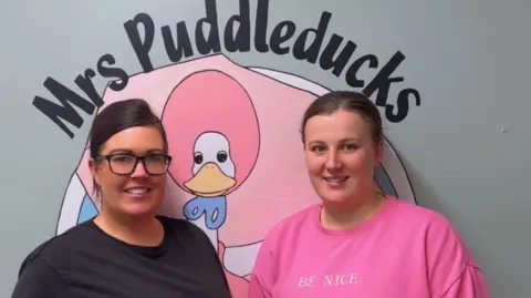 Katie Underwood Two women stood side by side, one with a pink t-shirt and the other in a pink jumper. Behind them the wall art reads 'Mrs Puddleducks'.