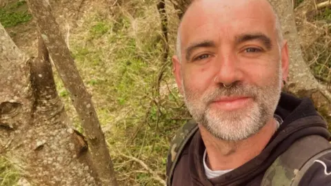 Stuart Loak Selfie of a man with a white and grey stubble beard and moustache with two nose rings.  He is standing in woodland, wearing a dark hoodie and smiling at the camera.