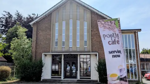 Acomb Methodist Church Exterior of a brick Methodist church with long vertical windows above the doorway. Outside the church is a sign advertising a pop up café. 