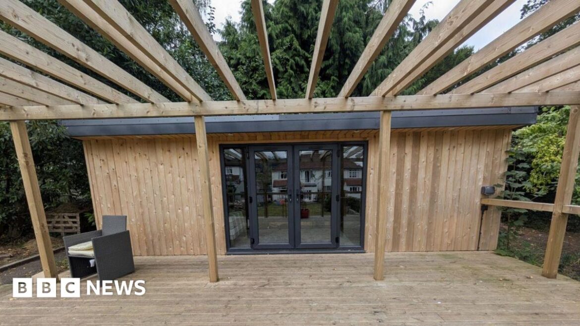 A wooden shed with a deck that has two glass doors and window next to each side of the door. A reflection shows a house on the opposite side and parts of the garden. There is a single grey chair in the corner of the shed.