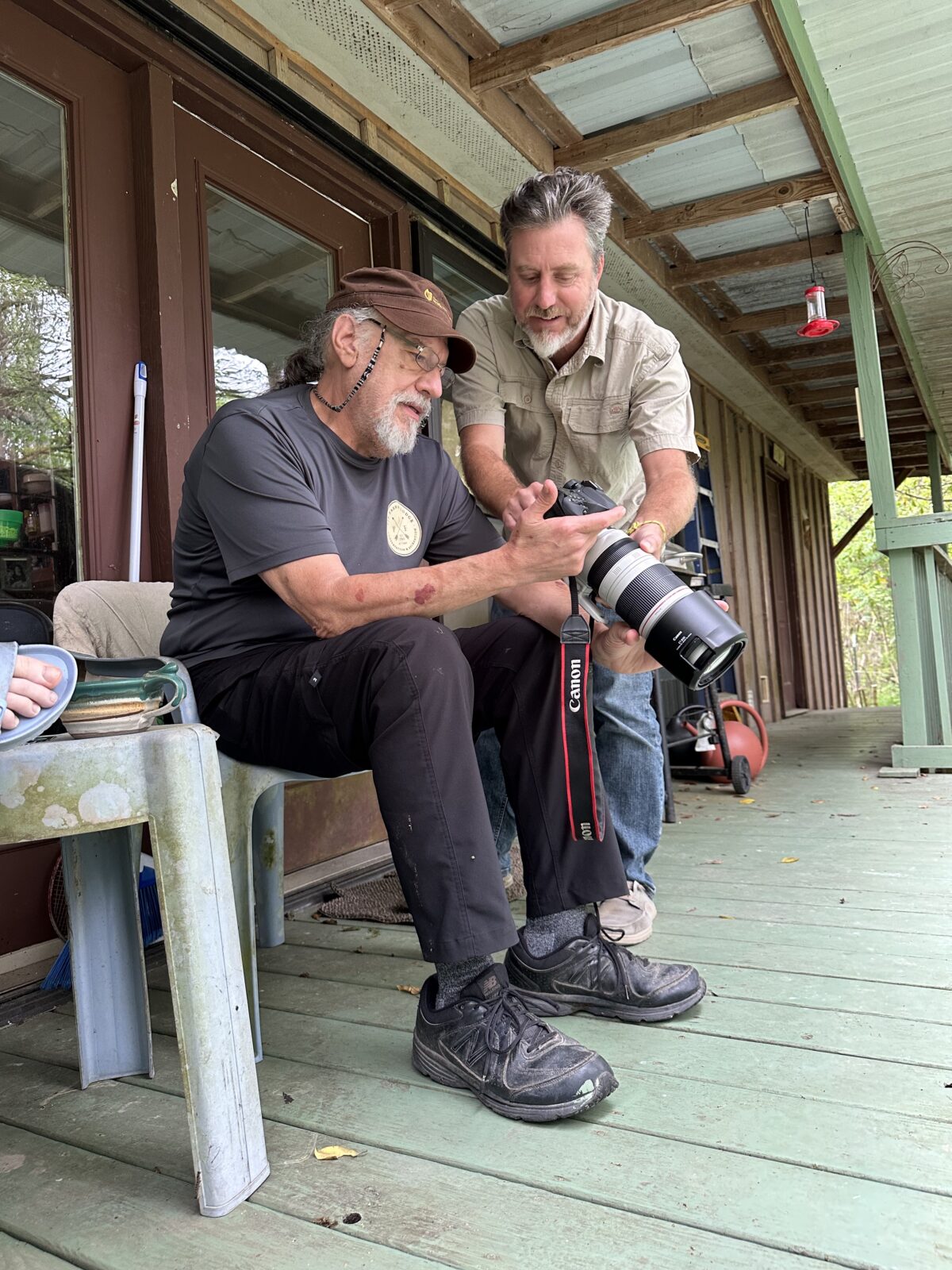 Two men on a porch look at a picture on a camera