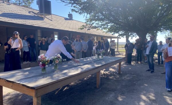 A person reaches for a lone glass of champagne on a long wooden table while a crowd holding glasses looks on.
