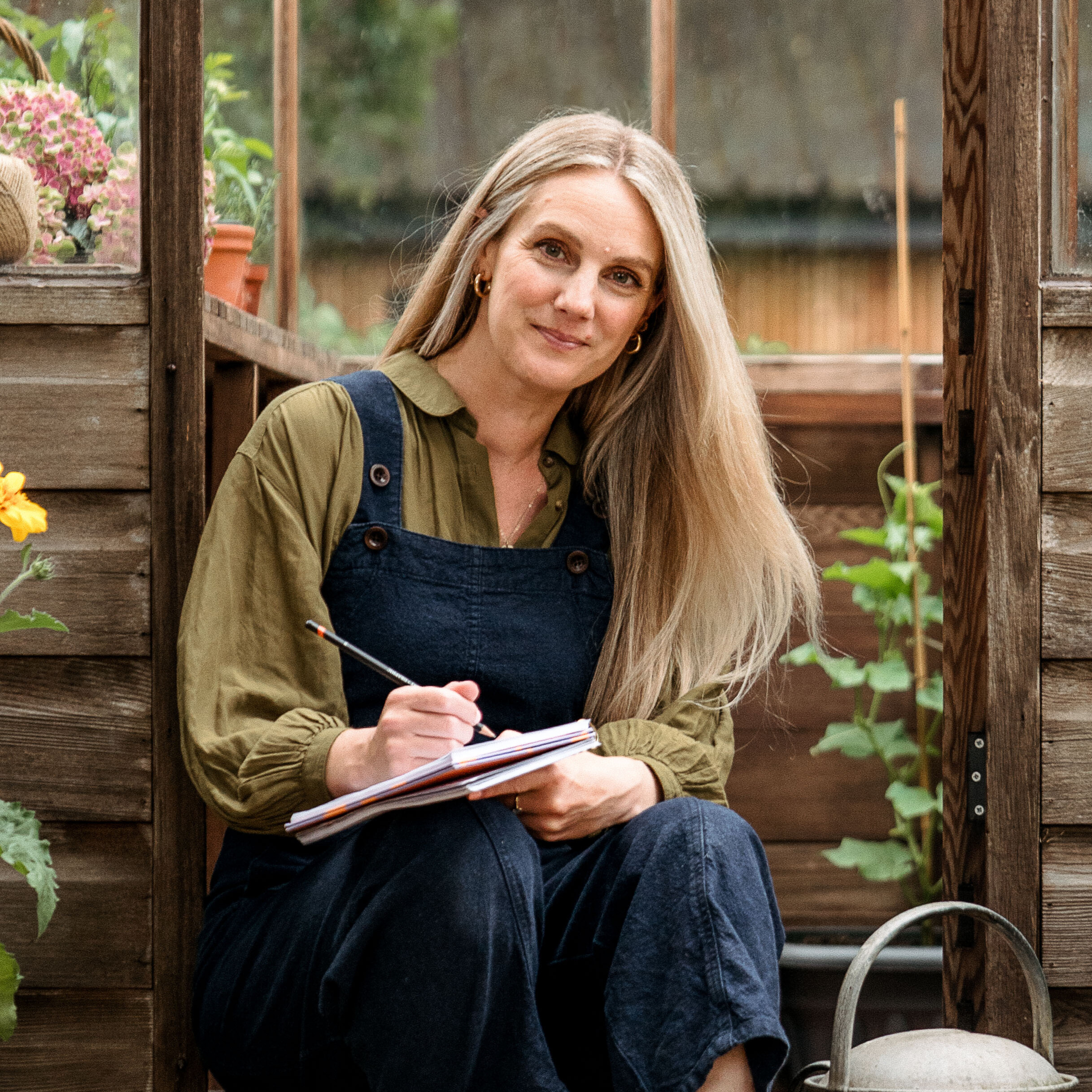 Woman sat on greenhouse step wearing navy dungarees and khaki shirt and holding a notebook
