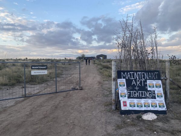 A sign reading "Maintenant Art + Fighting" next to an open fence gate, with a couple walking up a dusty road toward an open barn building.
