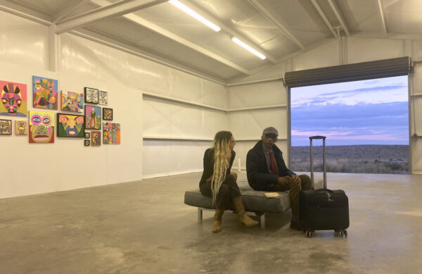 A person with long blond hair sits with an elderly man on a backless couch inside an art gallery, with paintings on one wall and an open barn door looking out on a desert landscape.