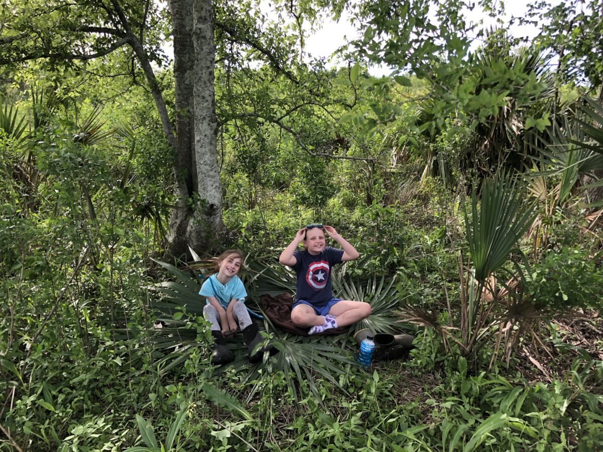 Two girls sit among wild growing fronds, trees and other plants