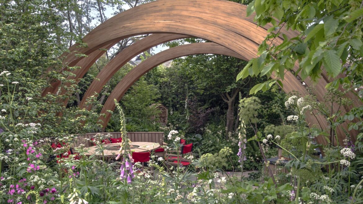 2026 landscape design trends: 5 experts say are here to stay A show garden at Chelsea Flower Show with trees, shrubs, and flowering perennials under a large wooden arch