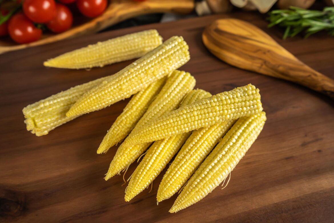 Harvest baby corn from your own garden this summer Several pieces of fresh baby corn are arranged on a wooden surface with cooking utensils and vegetables in the background.
