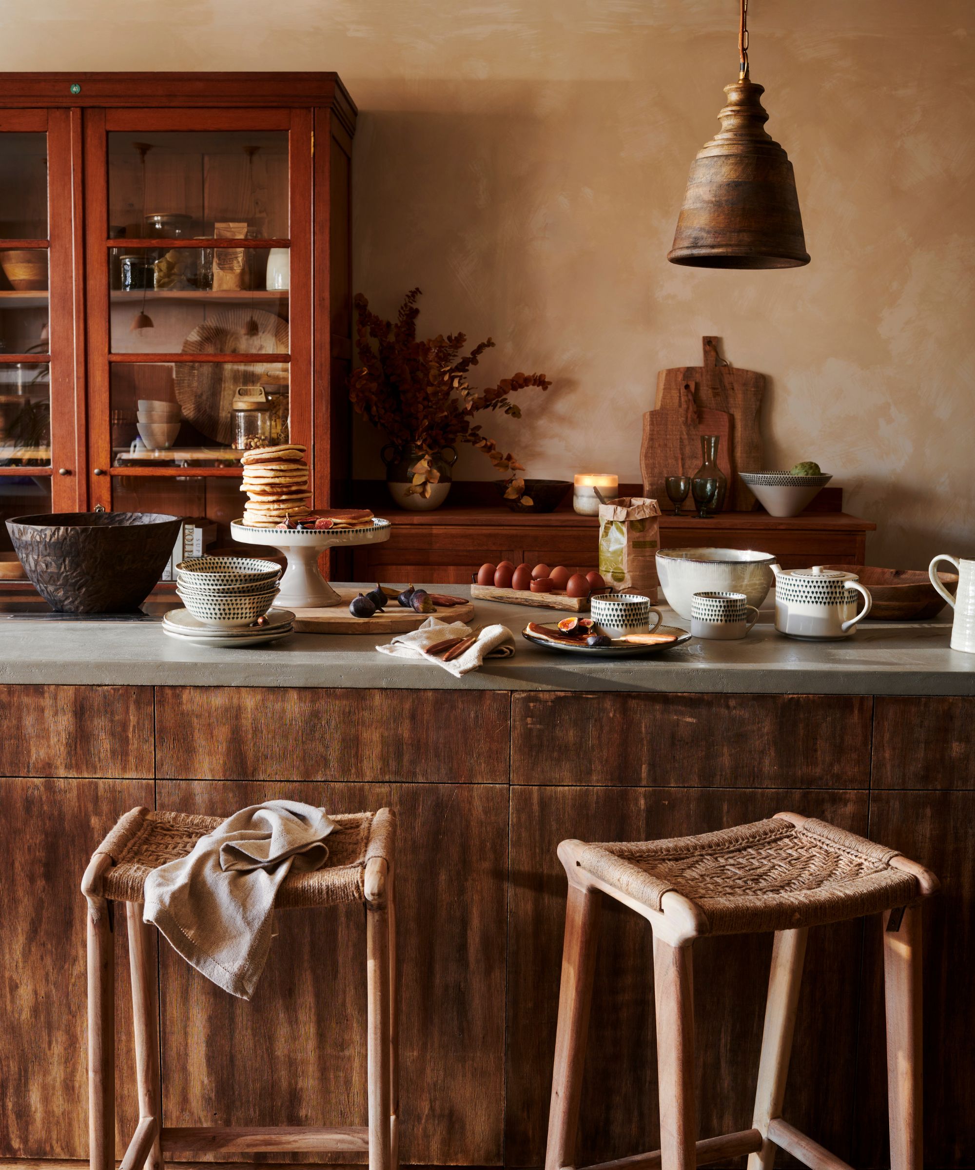 An earth toned kitchen, with wooden island and red painted pantry. Pancakes are stacked on a cake stand on the island. Two rattan stools pulled up.