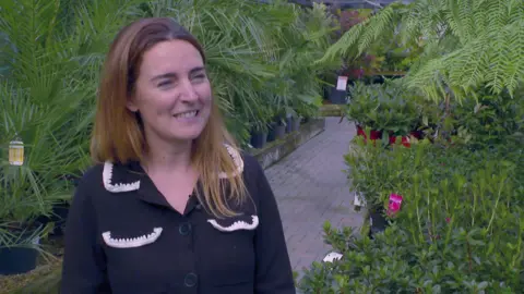 Amy Stewart smiles during a TV interview. She has long fair hair and is wearing a black blouse with white lace trim on the collar and breast pockets. She is standing among large palms and ferns in the plant sales area of the garden centre.