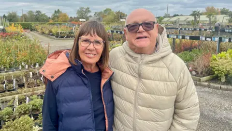 BBC A woman and a man are stood in the outside area of a garden centre. There are rows of plants and shrubs of different varieties. The man is bald and wearing sunglasses and a cream puffer coat. The woman is wearing glasses, a black long sleeve top, navy bodywarmer and had dark short hair. 
