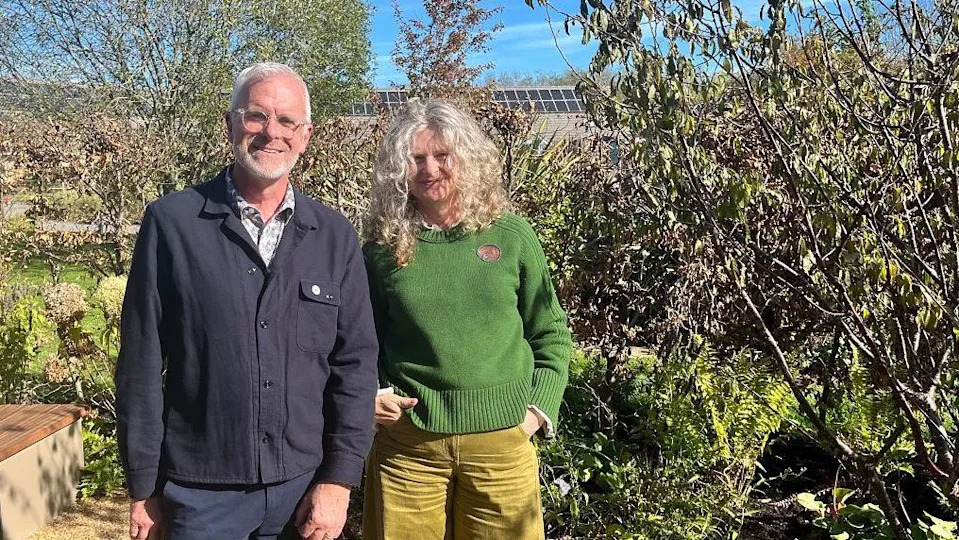 A man and a woman stand next to each other in a garden. The man is wearing a dark blue shirt and trousers, and the woman is wearing a dark green jumper with light green trousers. 