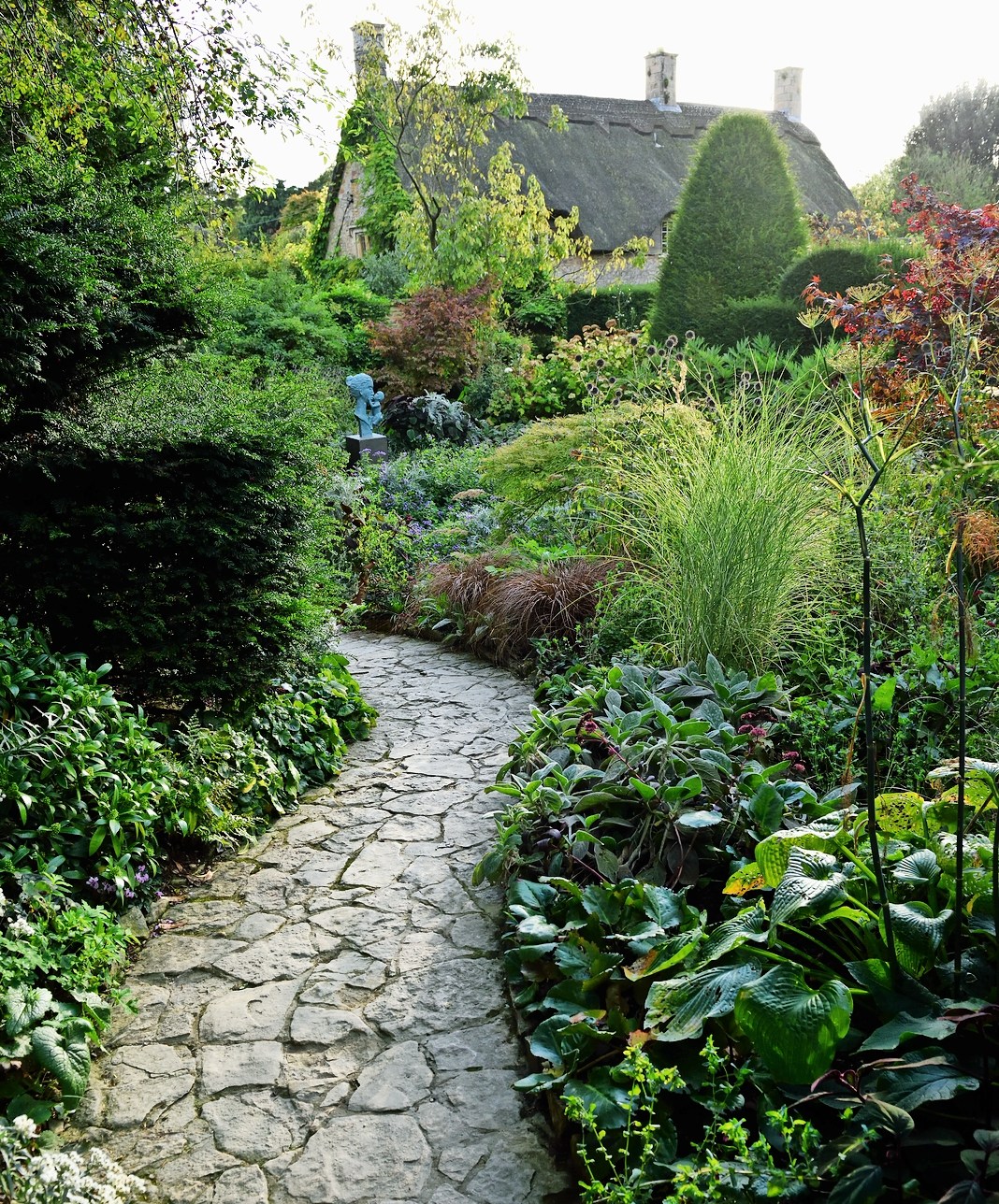 A stone path curves through the Hidcote Arts and Crafts garden, leading towards a thatched-roof house.