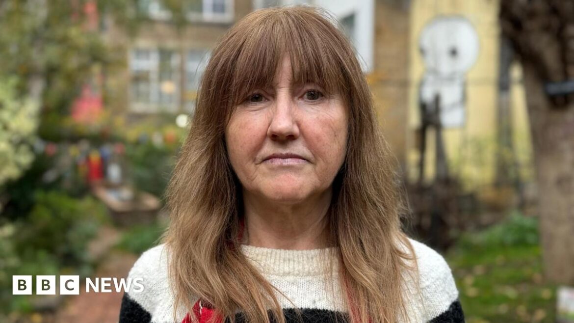 Louise Gates in black and cream striped jumper. She has brown hair past her shoulders and a fringe. She stands in the community garden, which is blurred behind her.
