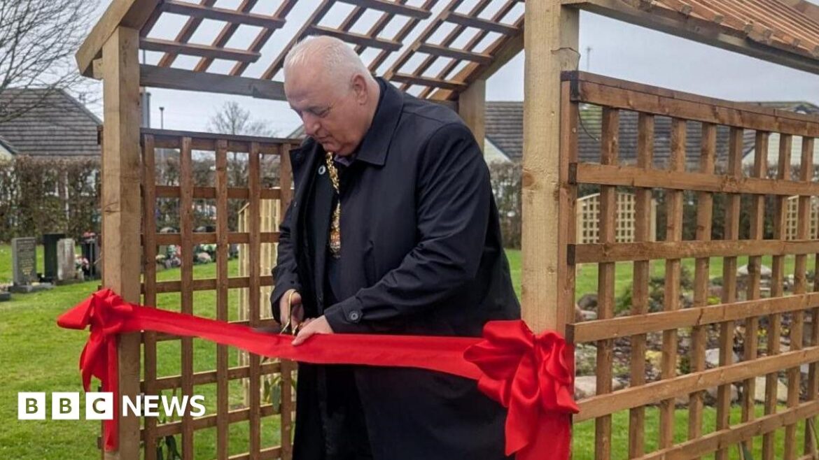 New pet garden officially opens at Carmountside Crematorium Lord Mayor of Stoke-on-Trent, Steve Watkins is stood under a wooden trellis with a red ribbon attached. He is holding a pair of scissors, and is cutting the ribbon.