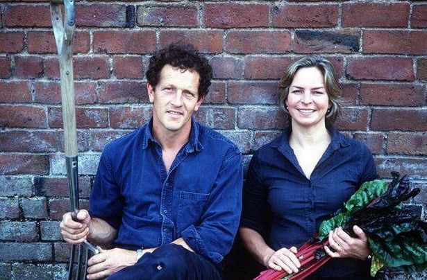 A picture of the couple wearing dark blue, Monty holding a trowel and Sarah some vegetables