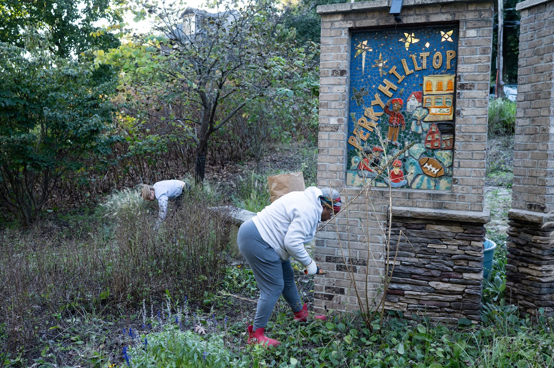 Two people in casual clothing work on weeding and cleaning up a garden area next to a brick sign that reads “Perry Hilltop.”.
