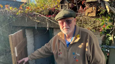 John Devine/BBC Colin Andrews has short grey hair and beard and has a green flat cap on his head. He has glasses with tinted lenses and is wearing a faun fleece over a blue, black and white shirt, he is holding the wooden door to the shelter open.