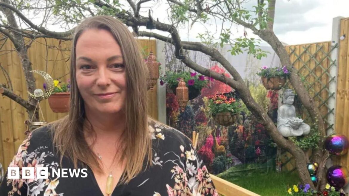 A woman with black floral top and long brown hair in a garden with wooden fences all around, a tree half way down and flowers at the back