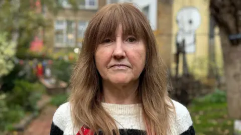 BBC Louise Gates in black and cream striped jumper. She has brown hair past her shoulders and a fringe. She stands in the community garden, which is blurred behind her. 