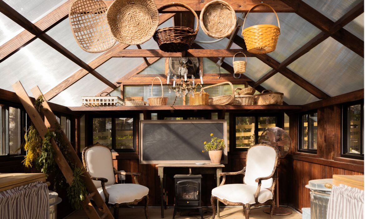 How to transform a high-functioning greenhouse into a cosy family sanctuary Greenhouse styled with antique chairs, a wood burning stove and wicker harvest baskets hanging from ceiling beam