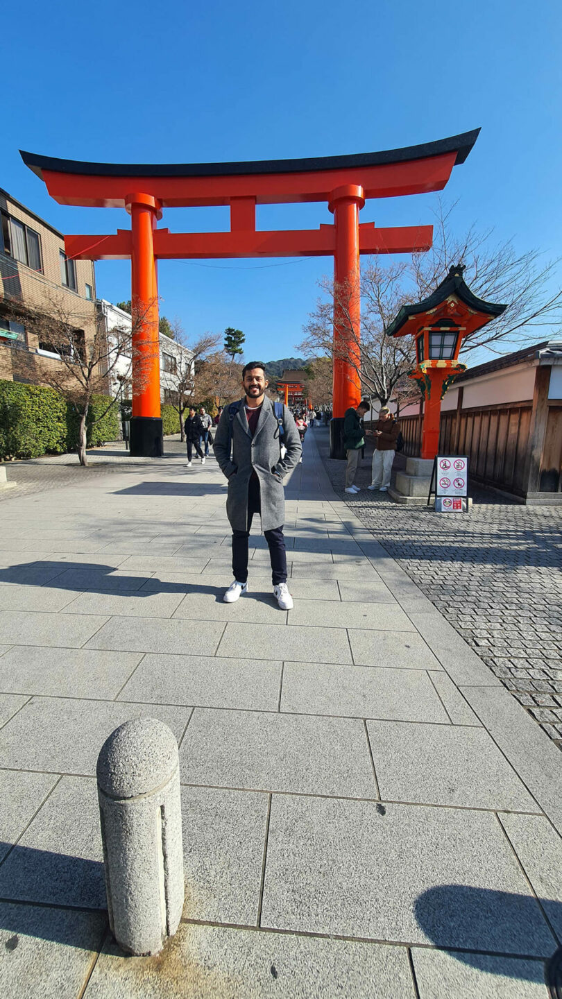 A person stands smiling in front of a large red torii gate on a sunny day, with a traditional lantern and several people visible in the background.