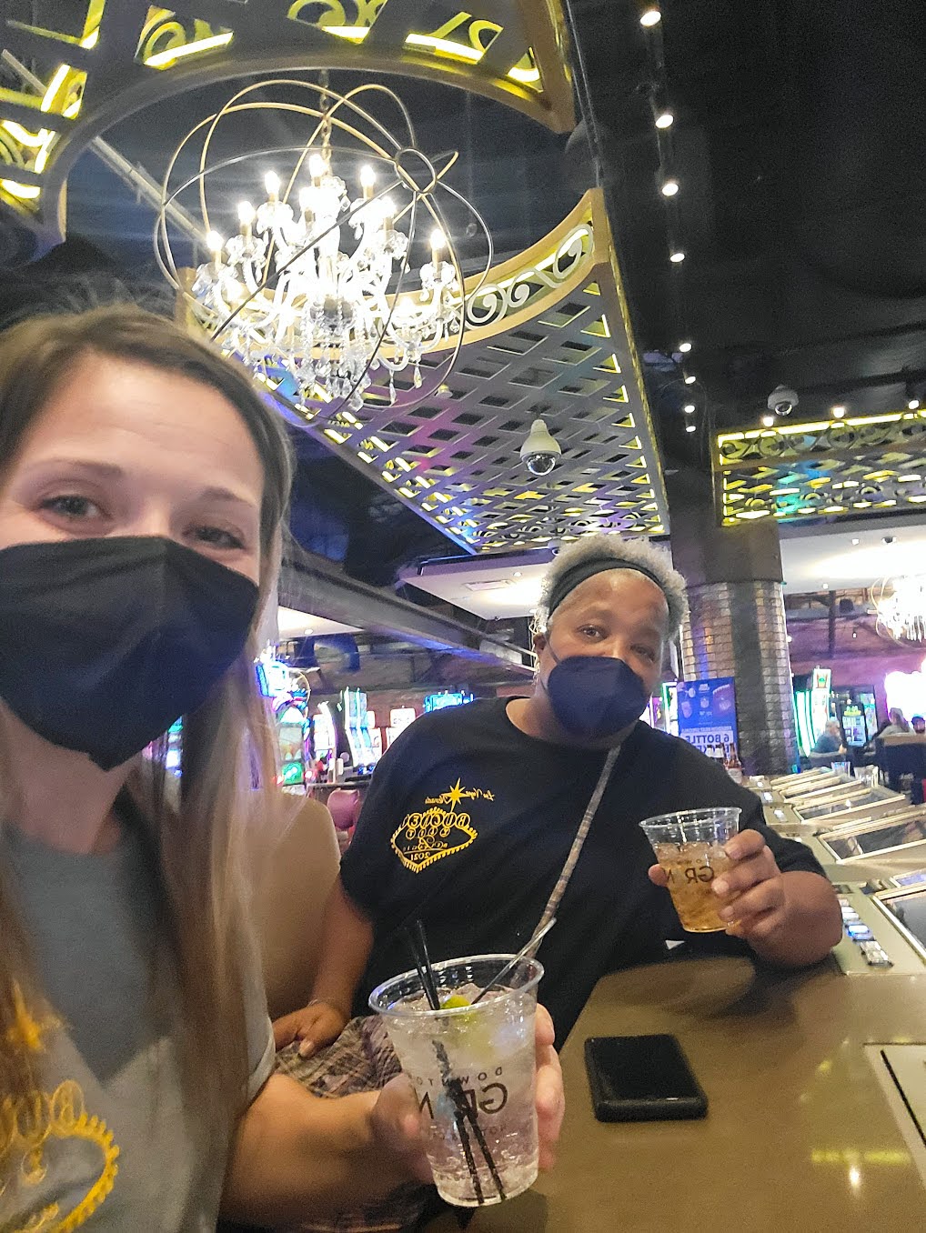 Two women wearing masks sit at a bar holding drinks, with empty glasses and a smartphone on the counter, under ornate lighting in a casino setting.