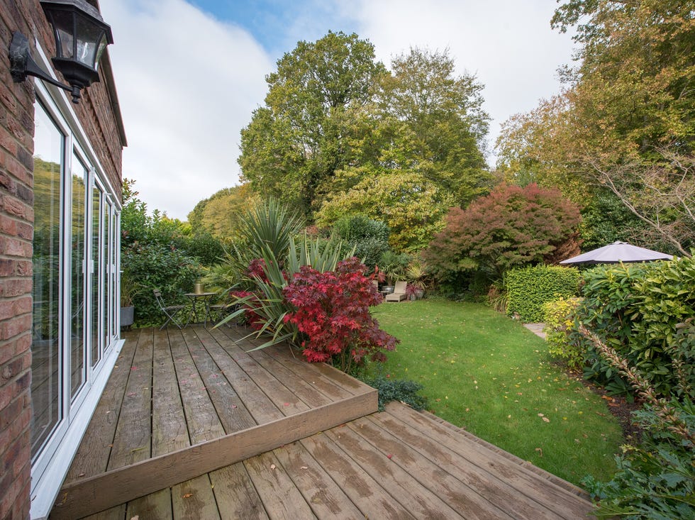 a general view of a house with bi fold / bifold patio doors wooden decking patio on to a back garden with two sun loungers, surrounded with trees, bushes and grass on a sunny day