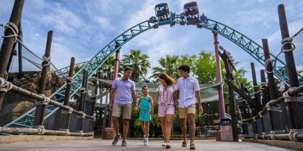 Busch Gardens Tampa Bay Condemned, Guests Left Devastated A group of four people, two men and two women, walk together through an amusement park. Behind them, a roller coaster with riders is visible against a blue sky. Palm trees and park decorations surround them.