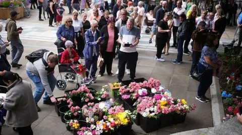 Getty Images Scores of people crowd around pots of flowers at a outdoor vigil to mark a year since the 2024 attack. The flowers are mainly pink, white and yellow.
