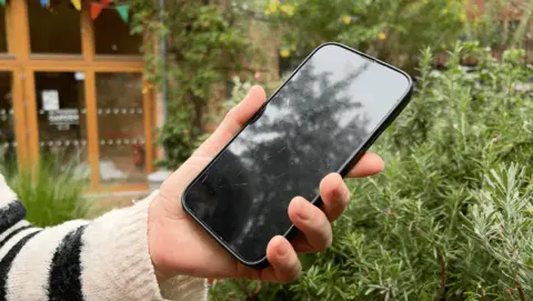 A woman wearing a black and cream striped jumper holds a smartphone in the community garden. 