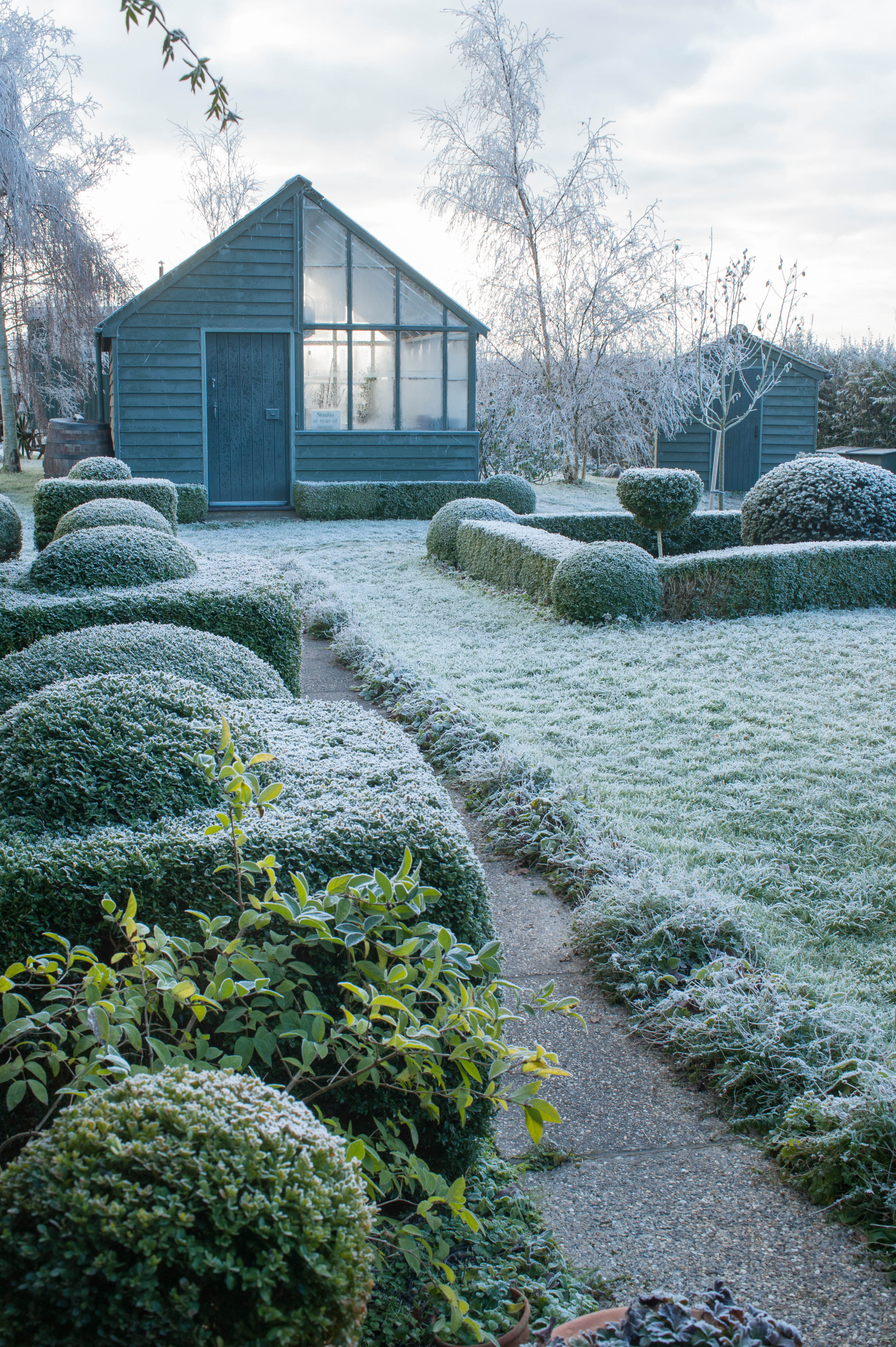 View of a frosted garden with a greenhouse, garden store, and sculpted hedges.