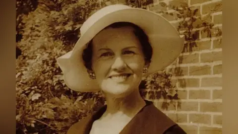 McKay family A headshot photo of Muriel McKay standing outdoors with a wall behind her that is partially covered in the creeper wisteria. She wears a flattering white rimmed hat, cluster earrings and a dark coloured coat. The photo is sepia coloured.