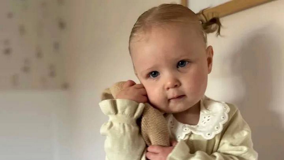 An infant is standing against a white wall, holding a plush toy in both hands. The baby has a serious expression and is dressed in a pale yellow outfit with ruffled sleeves.
