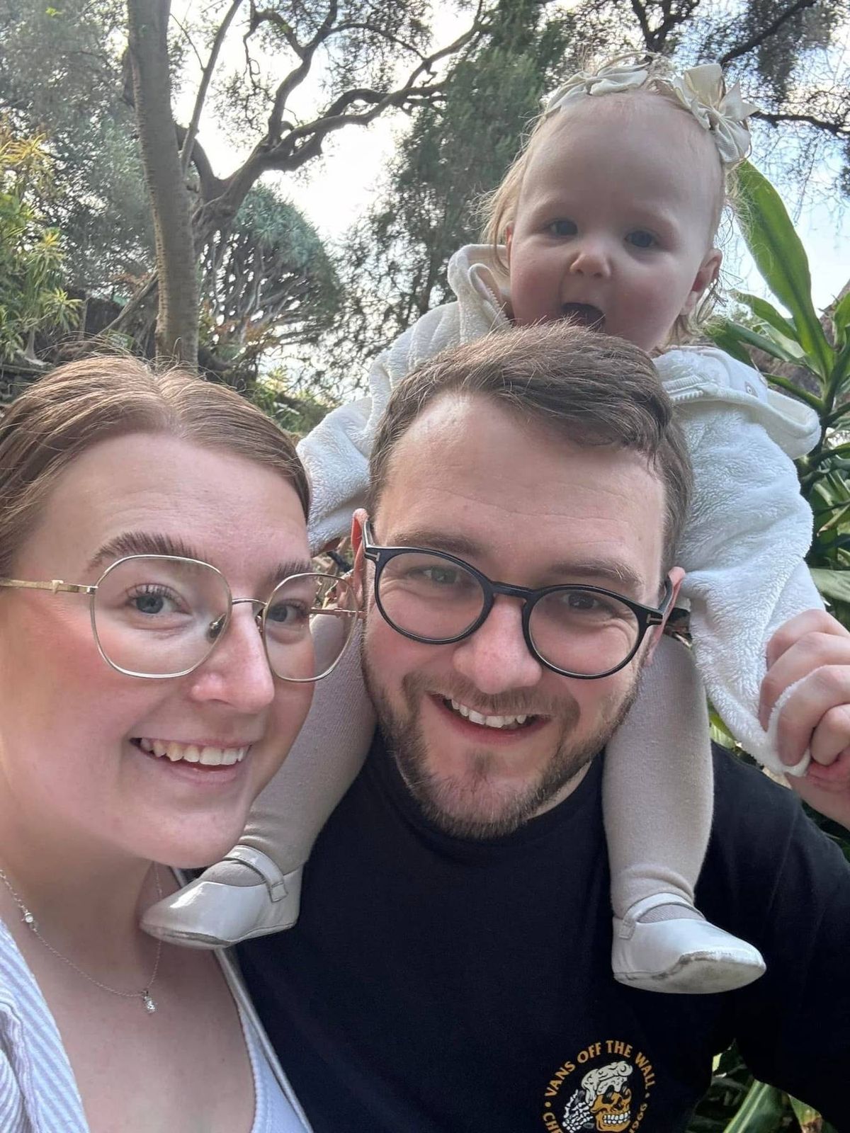 Two individuals wearing glasses are posing for a photograph with a child on one of their shoulders, all of them smiling in a garden setting.